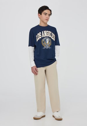 Granatowy T-shirt z napisem "LOS ANGELES" w kolorze kremowym, noszony na białej koszulce z długim rękawem; w połączeniu z beżowymi spodniami i białymi trampkami.