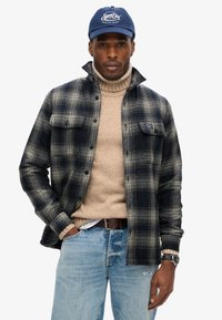 Mønstret skjorte i marineblå og creme, strikket tan turtleneck under, blå jeans, brun bælte og marineblå cap med logo. Flydende tekstur og lagdelt design.