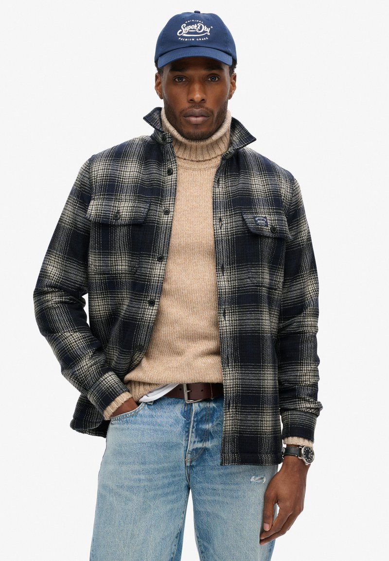 Mønstret skjorte i marineblå og creme, strikket tan turtleneck under, blå jeans, brun bælte og marineblå cap med logo. Flydende tekstur og lagdelt design.