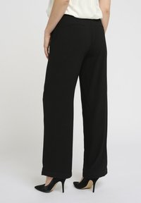 Zwarte losvallende broek met een gladde textuur, voorzien van een eenvoudig ontwerp, twee achterzakken en gedragen met puntige zwarte hiel.