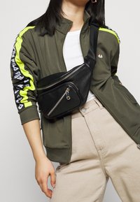 Czarna skórzana nerka z dwiema zamykanymi na zamek przegrodami, noszona na ciele. Posiada matowe wykończenie i regulowany pasek.