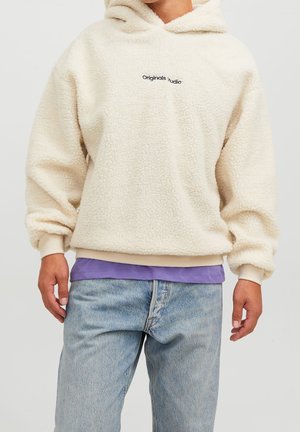 Person trägt einen cremefarbenen Sherpa-Hoodie mit dem Text "Originals Studio", darüber ein violettes Shirt und hellblaue Jeans.