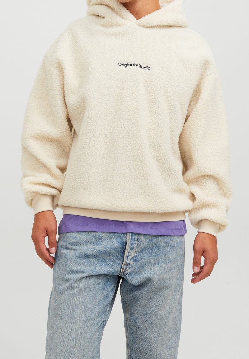 Personne portant un sweat à capuche sherpa de couleur crème avec le texte "Originals Studio", superposé à une chemise violette et un jean bleu clair.