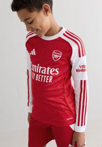 Maglia da calcio rossa con maniche lunghe bianche, caratterizzata da un motivo testurizzato, logo dell'Arsenal e testo "Emirates Fly Better". Inclusi pantaloni corti rossi.