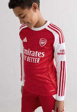 Rode voetbalshirt met lange witte mouwen, voorzien van een gestructureerd patroon, het Arsenal-logo en de tekst "Emirates Fly Better". Rode shorts inbegrepen.