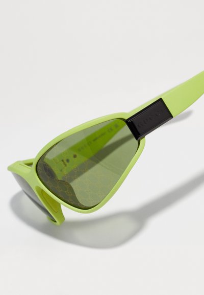 Gafas de sol de plástico verde con forma de lente triangular, tinte verde oscuro y un acento negro de marca en la patilla. Patrón de logo sutil en el interior.