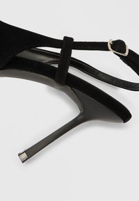 Fekete velúr tűsarkú szandál vékony bokapánttal, hegyes orral és fényes fémcsat díszítéssel. Magas sarok sima felülettel.