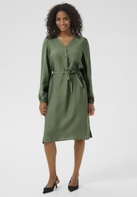 Une robe à manches longues, décolletée en V, de couleur vert olive, fabriquée dans un matériau lisse, avec un devant boutonné, une taille à nouer et des fentes sur les côtés.