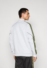 Homme portant une veste de survêtement blanche avec des rayures vertes et noires sur les manches, associée à un pantalon sombre, sur fond uni.
