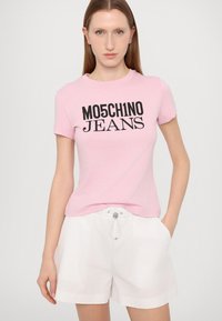 Ružová bavlnená tričko s čiernym textom "MO5CH1NO JEANS", spárované s bielymi šortkami na šnúrku. Jednoduchý dizajn s klasickým strihom.