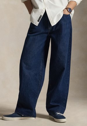 Personne portant un jean bleu foncé à jambes larges, des baskets bleues avec semelles blanches, une chemise blanche et une montre-bracelet au cadran bleu.