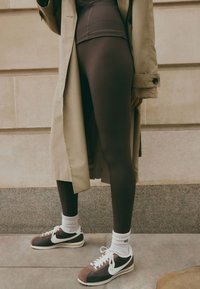 Ruskeat leggingsit yhdistettynä tumman sävyiseen trenssitakkiin ja ruskeisiin Nike-tennareihin. Tennareissa on valkoisia yksityiskohtia ja tekstuurillista kangasta.