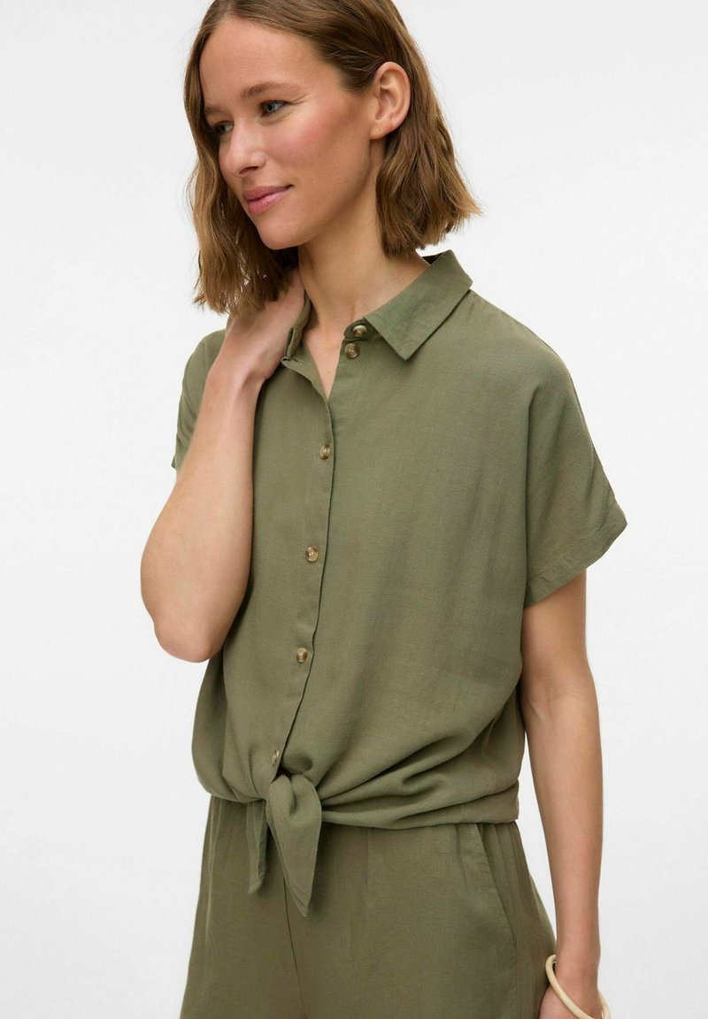 Camicia color verde oliva a maniche corte con colletto, chiusura a bottoni e design annodato in vita, realizzata in un tessuto leggero.