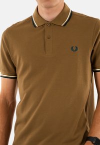 Bruine polo met een gestructureerde stof, voorzien van een drieknoopsluiting, een blauw embleem en contrasterende gestreepte trims op de kraag en mouwen.