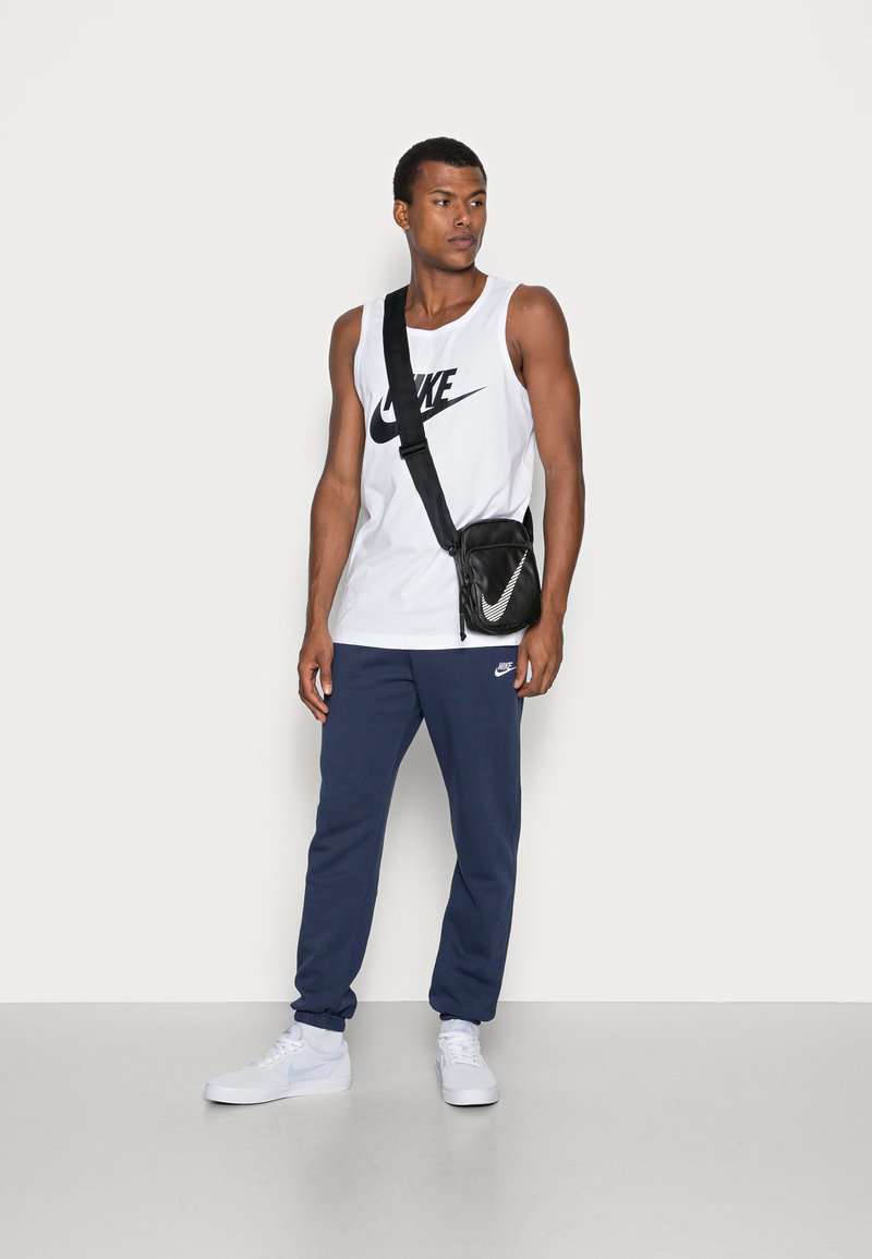 Débardeur Nike blanc avec logo noir, pantalon de survêtement bleu marine avec logo, sac bandoulière noir avec accent réfléchissant, et baskets blanches.