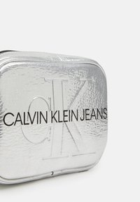 Stříbrná obdélníková kosmetická taštička s texturovaným povrchem, se zlaceným logem "CK" a nápisem "CALVIN KLEIN JEANS" v černém písmu. Zapínání na zip.