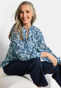 Leicht fallende Bluse in Blau und Weiß mit einem Schlüssel-Ausschnitt und langen Ärmeln, kombiniert mit dunklen Jeans, vor einem hellen Hintergrund.