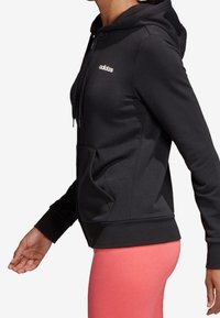 Sweat à capuche noir zippé en tissu doux avec poche avant et capuche à cordon, arborant un petit logo blanc sur la poitrine gauche.