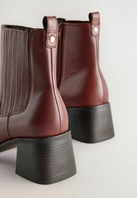 Botines de cuero burdeos con ribetes verticales, cremalleras laterales y tacones bloques negros. Textura lisa con costuras decorativas y detalles en dorado.
