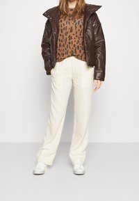 Braune Kunstleder-Puffermantel, Pullover mit Leopardenmuster, crèmefarbene weite Hose und weiße Sneaker, vor einem neutralen Hintergrund präsentiert.