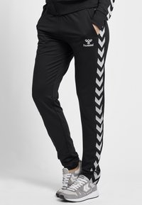 Svarta träningsbyxor med slim fit, utrustade med ett vitt chevronrand längs sidan och Hummel-logotypen fram. Tillverkade av lätt material.