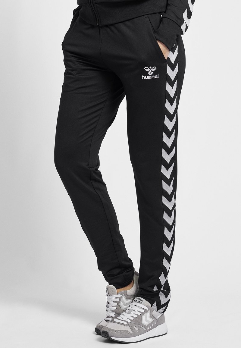 Svarta träningsbyxor med slim fit, utrustade med ett vitt chevronrand längs sidan och Hummel-logotypen fram. Tillverkade av lätt material.