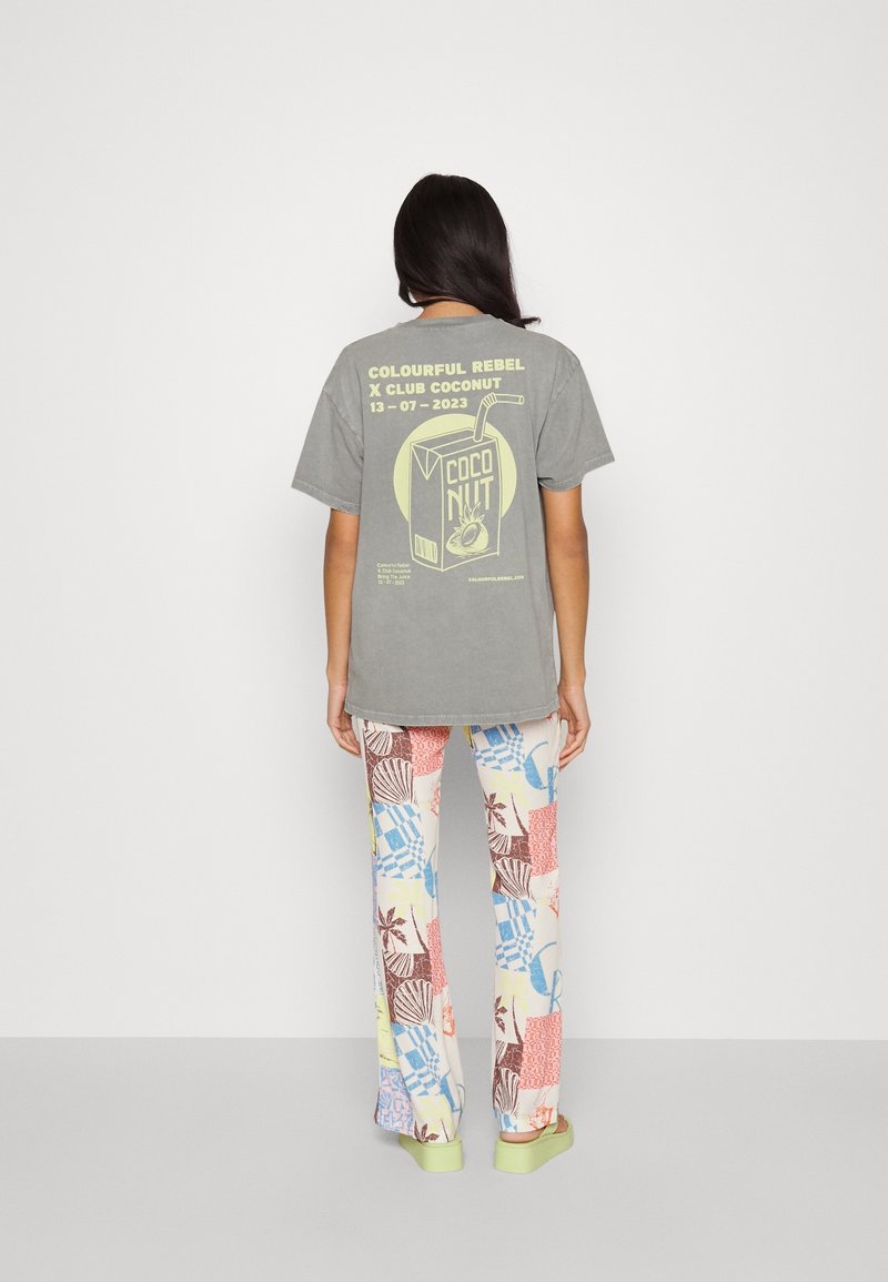 Grå oversized t-shirt med tryckt design av kokosnötskartong; kombinerad med färgglada, mönstrade byxor med olika tropiska motiv. Gröna sandaler.