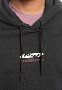 Felpa con cappuccio grigio scuro con coulisse, un piccolo logo bianco e rosa, e una texture morbida. Indossata sopra una maglietta rosa chiaro.