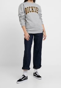 Sweat-shirt gris avec l'inscription "DICKIES" en lettres jaunes, associé à un pantalon rollé à rayures fines foncé et des baskets noires avec des accents blancs.