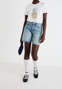 Vit t-shirt med en ananasgrafik, ljusblå denimshorts, knälånga vita strumpor och svarta Mary Jane-skor, bärande en marinblå väska.