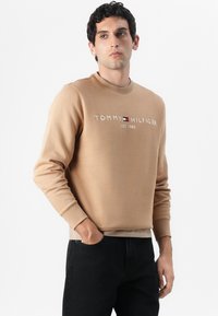 Lysebrun sweatshirt med rund hals og broderet "TOMMY HILFIGER EST. 1985" logo. Parret med mørke jeans i slim fit.