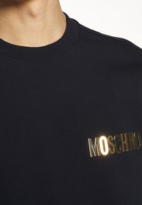 Černá mikina s žebrovaným výstřihem, s zlatým logem Moschino přes hruď. Jemný materiál s hladkou texturou.