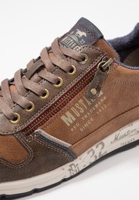 Sneaker en cuir marron avec une finition texturée, des accents contrastants, des éléments en or, et un détail de fermeture éclair. Présente un logo dans un style vintage.