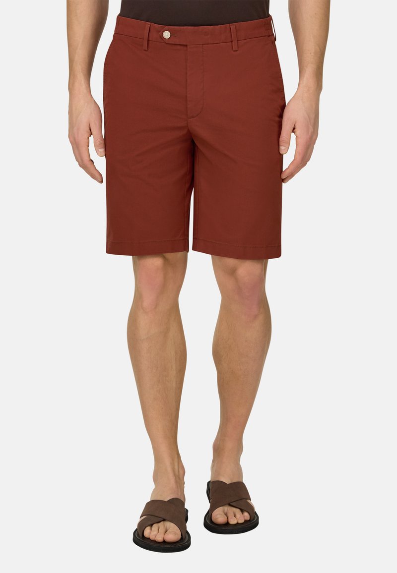 Homme portant un short couleur rouille et des sandales marron entrecroisées, debout devant un fond clair uni.