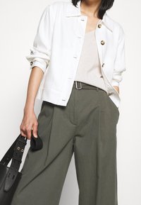 White cotton jacket with gold buttons, cream top underneath, and green high-waisted trousers. Holding a black handbag with a textured finish.