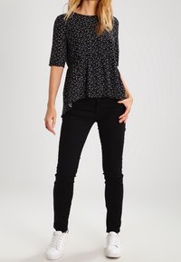 Blusa nera a peplum con pois bianchi, abbinata a jeans neri slim e sneakers bianche. Il tessuto sembra morbido con una vestibilità comoda.