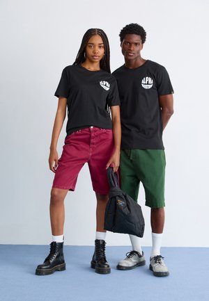 Two models posed together: one in a black graphic tee and maroon shorts, the other in a black tee and green shorts. Both wear boots.