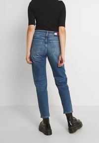 Person wearing high-waisted blue jeans with back pockets and black ankle boots, standing against a plain white background.