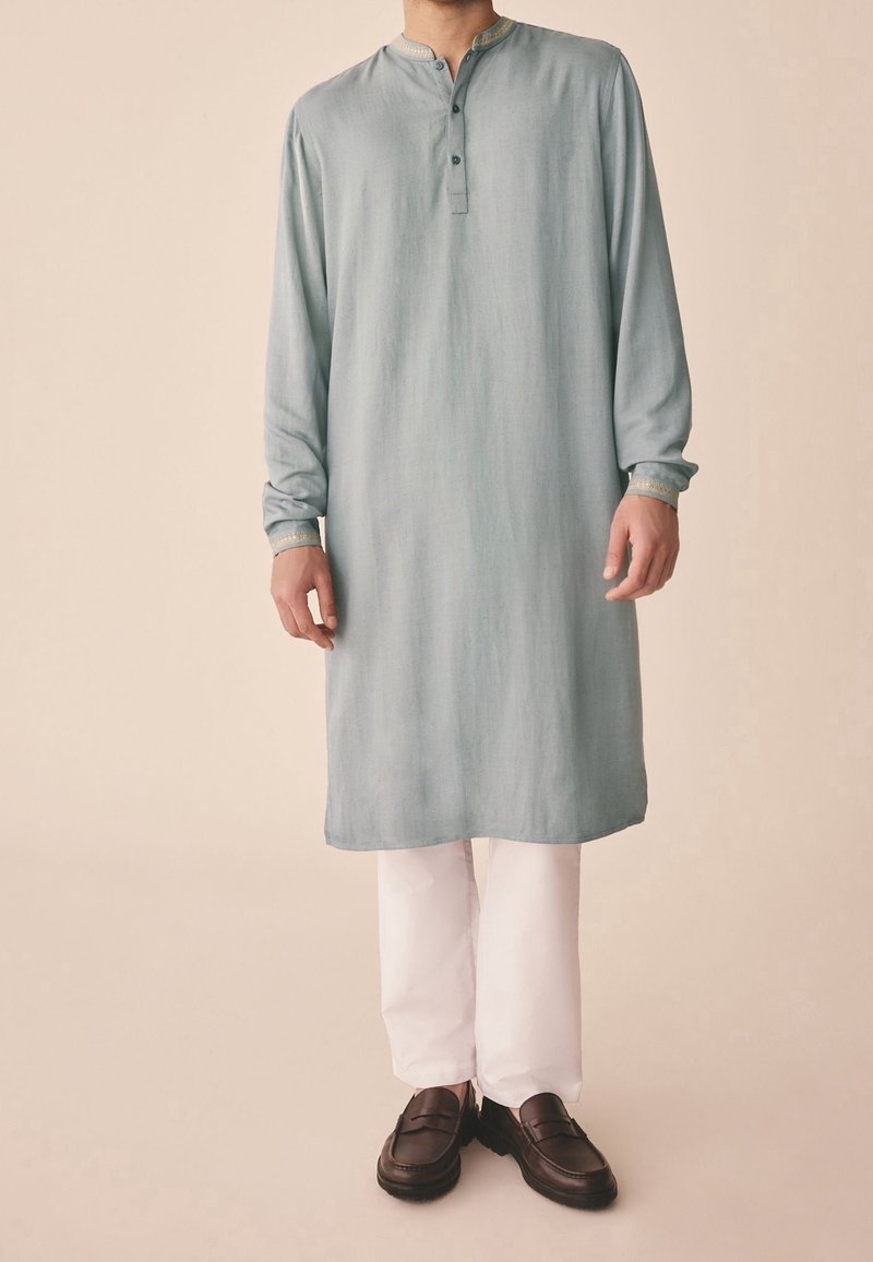 Homme portant une tunique bleu clair mi-genoux avec col mandarin, pantalon blanc et mocassins marron foncé sur fond beige.