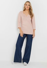 Maglietta rosa chiaro a maniche lunghe con scollatura a V e pieghe frontali, abbinata a jeans a zampa d'elefante blu scuro e sneakers bianche.
