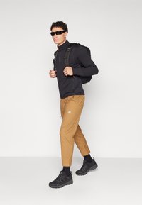 Black zip-up jacket, tan elastic pants with logo, black sneakers, and sunglasses. Backpack featured with adjustable straps and pockets.