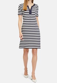 Woman wearing knee-length, short-sleeve navy and white horizontal striped dress with a navy V-neck collar, standing with white heels.