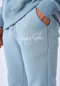 Pantaloni da tuta azzurro chiaro con una texture liscia, dotati di un logo ricamato "Project x Paris" in bianco nell'area della tasca.