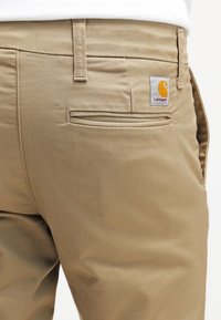 Khakibroek gemaakt van duurzaam materiaal, voorzien van een achterzak met een klein Carhartt-logo en een gladde textuur met subtiele stiksels.