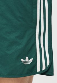 Πράσινα αθλητικά σορτς με τρεις λευκές ρίγες και λευκό λογότυπο trefoil της Adidas κοντά στο τελείωμα.