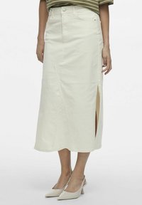 Falda midi de mezclilla blanca con una abertura frontal, con trabillas para cinturón y cierre de botón. Combinada con tacones puntiagudos de color claro.