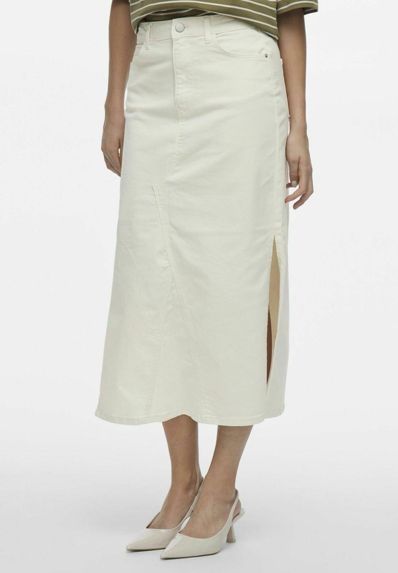 Falda midi de mezclilla blanca con una abertura frontal, con trabillas para cinturón y cierre de botón. Combinada con tacones puntiagudos de color claro.