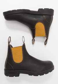 Botas chelsea de couro castanho com painéis elásticos amarelos, alças de puxar e solas de borracha preta robustas. O acabamento costurado melhora a durabilidade.