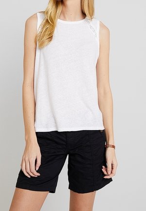 Valkoinen hihaton pusero, jossa pitsiyksityiskohtia olkapäillä, yhdistetty mustiin polvipituisiiin shortseihin. Asussa on rento istuvuus ja pehmeä kangas.