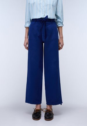 Wijde, marineblauwe broek gemaakt van een gladde stof, met een hoge taille, een strikceintuur en zijzakken. Het schoeisel is donker met veters.